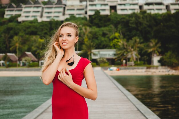 Junge schöne attraktive Frau, die allein auf Pier im Luxusresorthotel, Sommerferien, rotes langes Kleid, blondes Haar, sexy Kleidung, tropischer Strand, verführerisch, sinnlich, lächelnd steht