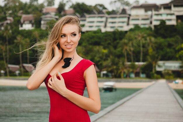 Junge schöne attraktive Frau, die allein auf Pier im Luxusresorthotel, Sommerferien, rotes langes Kleid, blondes Haar, sexy Kleidung, tropischer Strand, verführerisch, sinnlich, lächelnd steht