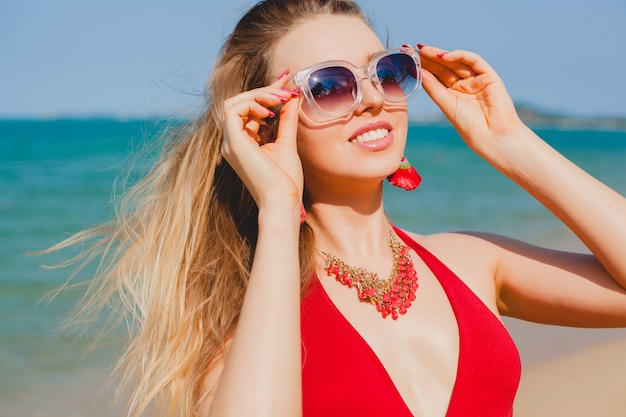 Junge schöne blonde Frau, die am Strand im roten Badeanzug, Sonnenbrille sonnenbad