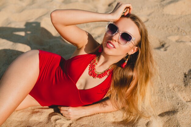 Junge schöne blonde Frau, die auf Sandstrand im roten Badeanzug, Sonnenbrille sonnenbad