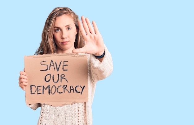 Kostenloses Foto junge schöne blonde frau hält mit offener hand das banner „rettet unsere demokratie“ und macht ein stoppschild mit ernster und selbstbewusster verteidigungsgeste