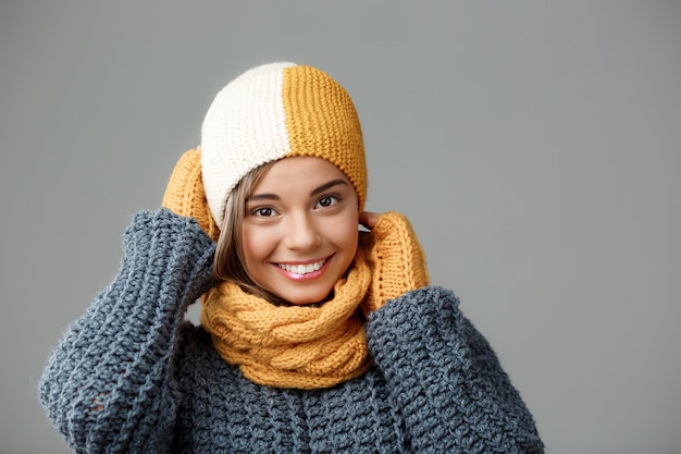 Junge schöne blonde Frau im Strickmützenpulloverschal und in den Handschuhen, die auf Grau lächeln.