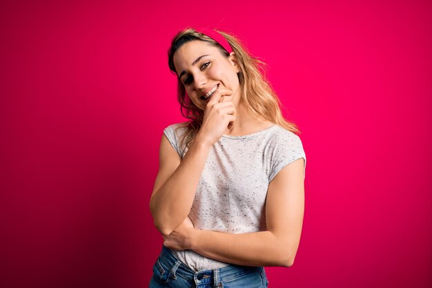 Junge schöne blonde Frau in lässigem T-Shirt steht vor isoliertem rosa Hintergrund und blickt selbstbewusst in die Kamera, lächelt mit verschränkten Armen und der Hand am Kinn und denkt positiv