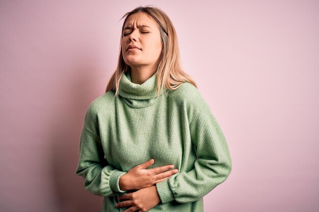 Junge schöne blonde Frau trägt Winterwollpullover über rosa isoliertem Hintergrund mit der Hand auf dem Bauch, weil Übelkeit schmerzhafte Krankheit sich unwohl fühlt Schmerzkonzept