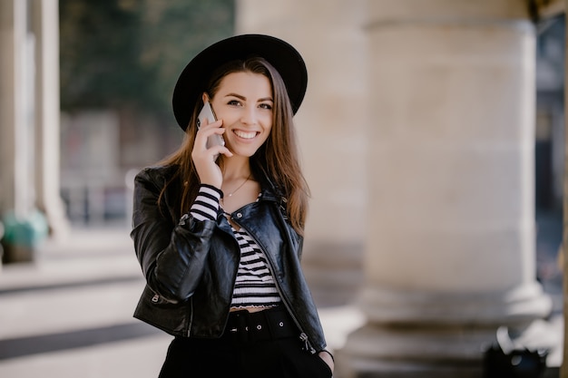 Junge schöne braunhaarige Mädchen in einer Lederjacke, schwarzer Hut auf der Stadtpromenade haben ein Telefongespräch