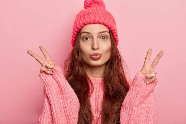 Kostenloses Foto junge schöne brünette europäische frau hält lippen gerundet, macht siegesfriedensgeste, trägt strickmütze und übergroßen pullover, hat langes haar, isoliert über rosa hintergrund.