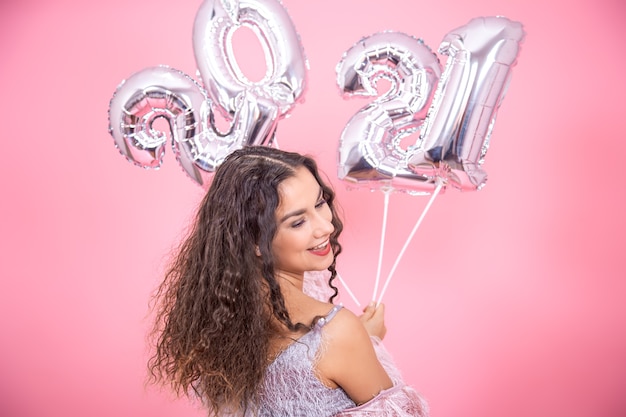 Junge schöne Brünette mit lockigem Haar und nackten Schultern, die auf einem rosa Hintergrund mit silbernen Luftballons für das Neujahrskonzept lächeln