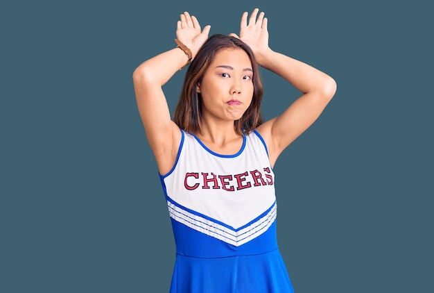 Junge schöne Chinesin in Cheerleader-Uniform macht Hasenohren-Gesten mit Handflächen und sieht zynisch und skeptisch aus