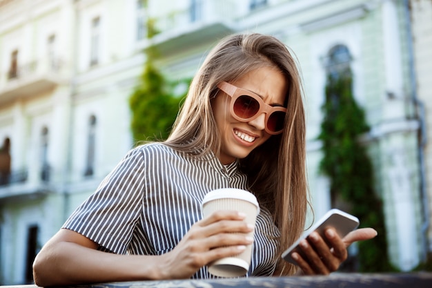 Kostenloses Foto junge schöne frau, die auf bank sitzt, telefon betrachtet, lächelnd.