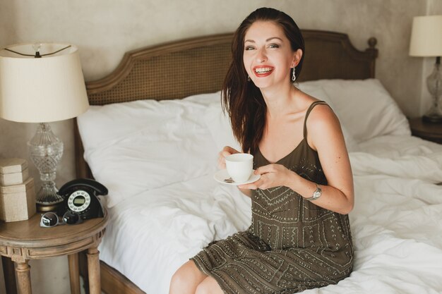 Junge schöne Frau, die auf Bett im Hotel, stilvolles Kleid, sinnliche Stimmung sitzt, Kaffee trinkt, Tasse hält