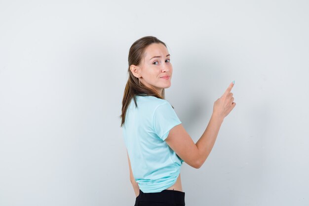 Junge schöne Frau, die auf die obere rechte Ecke im T-Shirt zeigt und fröhlich aussieht.