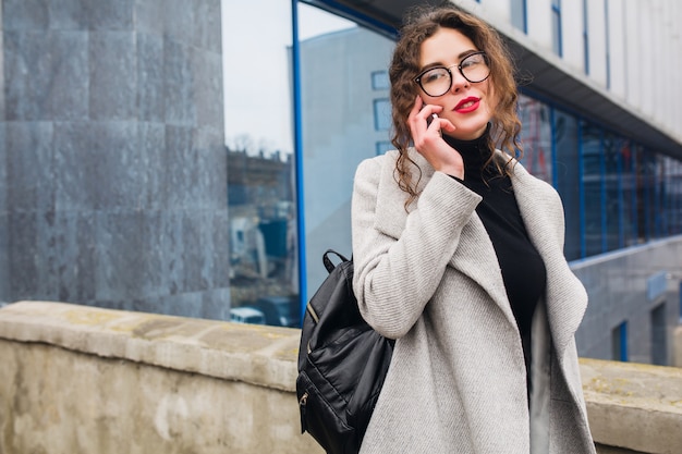 Junge schöne Frau, die auf Smartphone spricht, Herbststraßenstadtstil, warmer Mantel, Brille, glücklich, lächelnd, Telefon in der Hand haltend, lockiges Haar