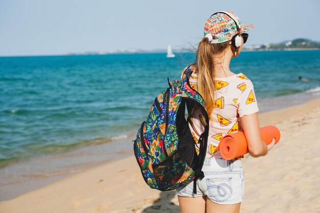 Junge schöne Frau, die auf Strand hält, die Yogamatte, Hipster-Sport-Beute-Stil, Jeansshorts, T-Shirt, Rucksack, sonnig, Sommerwochenende hält