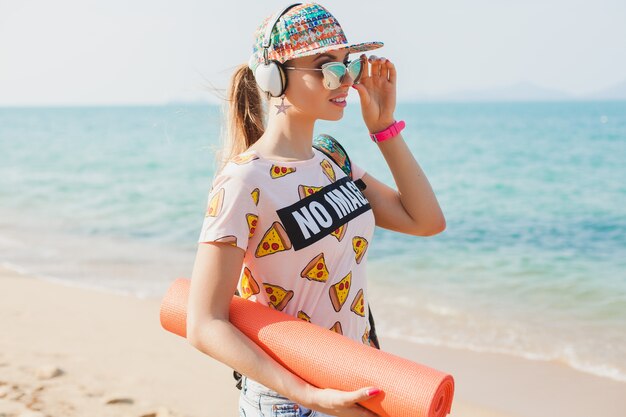 Junge schöne Frau, die auf Strand mit Yogamatte geht, Musik auf Kopfhörern, Hipster-Sport-Beute-Stil, Jeansshorts, T-Shirt, Rucksack, Mütze, Sonnenbrille, sonnig, Sommerwochenende, fröhlich hört