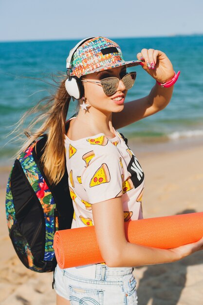 Junge schöne Frau, die auf Strand mit Yogamatte geht, Musik auf Kopfhörern, Hipster-Sport-Beute-Stil, Jeansshorts, T-Shirt, Rucksack, Mütze, Sonnenbrille, sonnig, Sommerwochenende, fröhlich hört