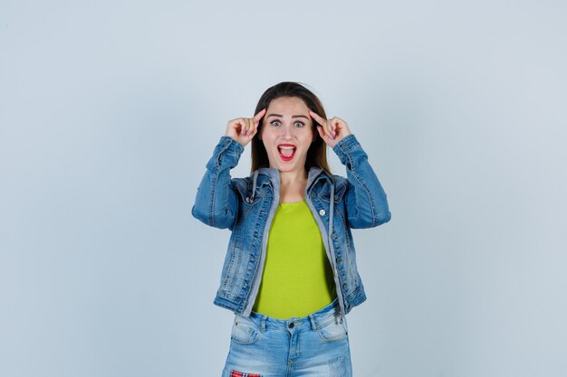 Junge schöne Frau, die Finger an den Schläfen in Denim-Outfit hält und verspielt aussieht, Vorderansicht.