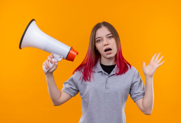 junge schöne Frau, die graues T-Shirt hält, das Lautsprecher auf isolierter gelber Wand hält