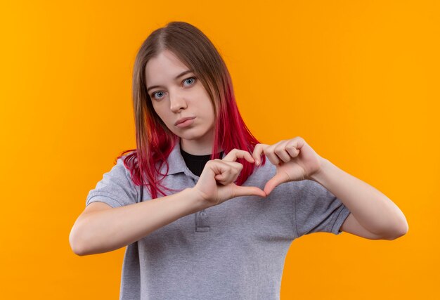 junge schöne Frau, die graues T-Shirt trägt, das Herzgeste auf isolierter gelber Wand zeigt