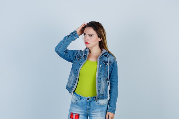 Junge schöne Frau, die Hand auf dem Kopf hält, im Denim-Outfit beiseite schaut und nachdenklich aussieht. Vorderansicht.