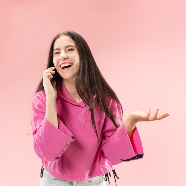 Junge schöne frau, die handy-studio auf rosa farbstudiohintergrund verwendet. konzept der menschlichen gesichtsgefühle. trendige farben