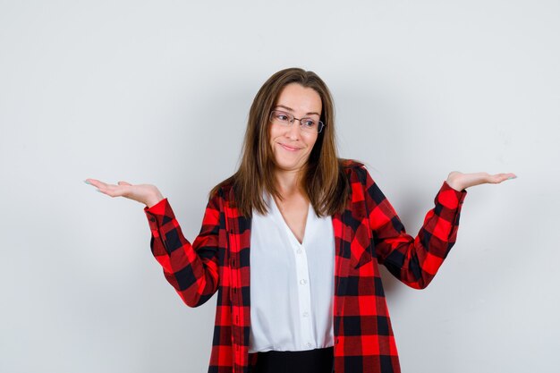 Junge schöne Frau, die hilflose Geste zeigt, in lässigem Outfit wegschaut und fröhlich aussieht, Vorderansicht.