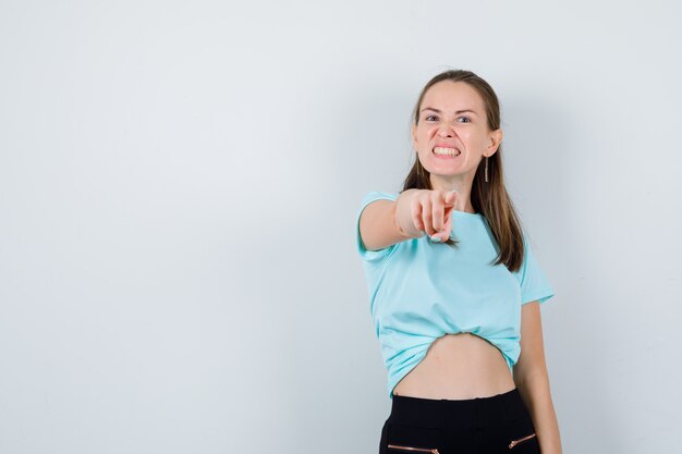 Junge schöne Frau, die im T-Shirt auf die Kamera zeigt und aggressiv aussieht. Vorderansicht.