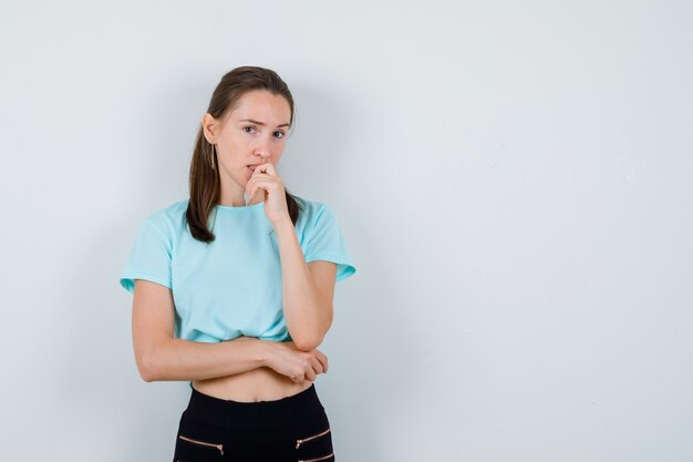 Junge schöne Frau, die in denkender Haltung im T-Shirt steht und verwirrt schaut, Vorderansicht.