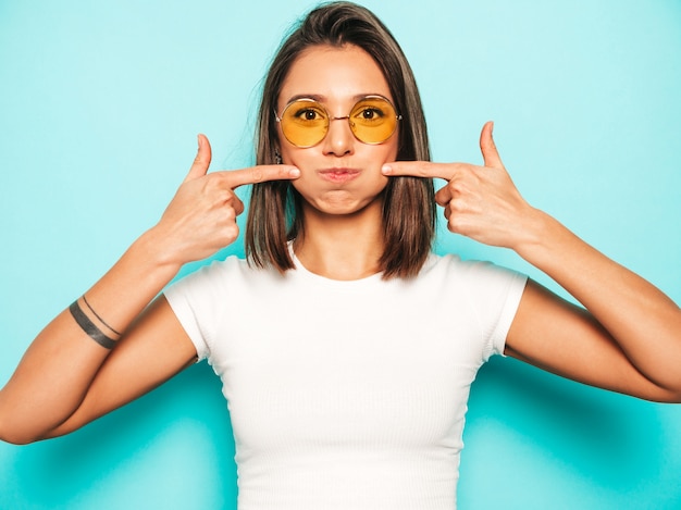 Junge schöne Frau, die Kamera betrachtet. Trendy Mädchen im lässigen weißen Sommer-T-Shirt und im gelben Rock in der runden Sonnenbrille. Positive Frau zeigt Gesichtsgefühle. Lustiges Modell, das ihre Wangen bläst.