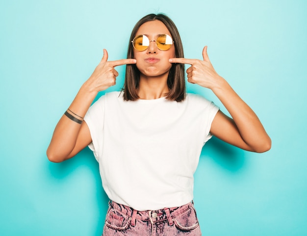 Junge schöne Frau, die Kamera betrachtet. Trendy Mädchen im lässigen weißen Sommer-T-Shirt und in den Jeansshorts in der runden Sonnenbrille. Positive Frau zeigt Gesichtsgefühle. Lustiges Modell, das ihre Wangen bläst.