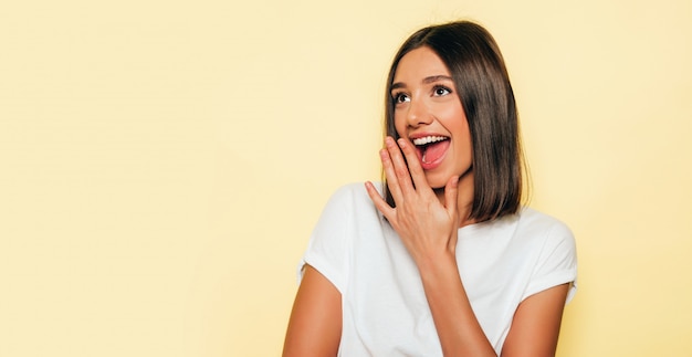 Junge schöne Frau, die Kamera betrachtet. Trendy Mädchen im lässigen weißen Sommer-T-Shirt und in den Jeansshorts. Positive Frau zeigt Gesichtsgefühle. Schockiert und überrascht mit Händen in der Nähe des Gesichts