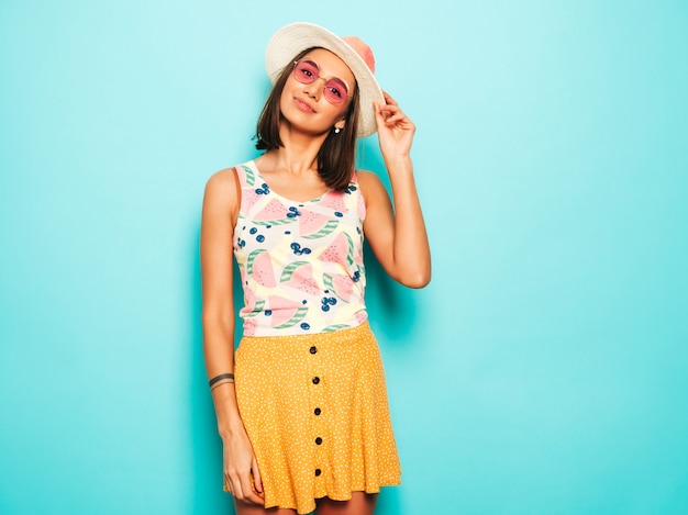 Junge schöne Frau, die Kamera im Hut betrachtet. Trendy Mädchen im lässigen weißen Sommer-T-Shirt und im gelben Rock in der runden Sonnenbrille. Positive Frau zeigt Gesichtsgefühle. Lustiges Modell lokalisiert auf Blau