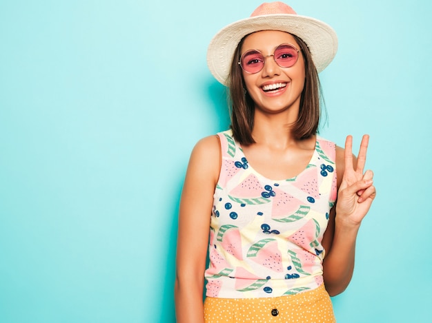 Junge schöne Frau, die Kamera im Hut betrachtet. Trendy Mädchen im lässigen weißen Sommer-T-Shirt und im gelben Rock in der runden Sonnenbrille. Positive Frau zeigt Gesichtsgefühle. Zeigt Friedenszeichen