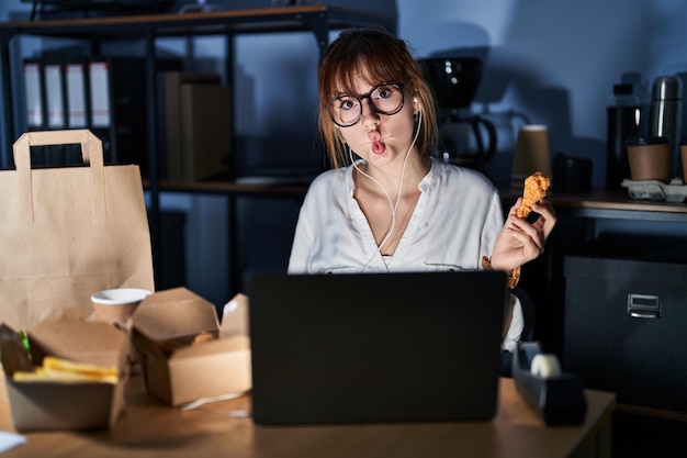 Kostenloses Foto junge schöne frau, die mit einem computer-laptop arbeitet und lieferlebensmittel isst, macht fischgesicht mit lippen, verrückte und komische geste. lustiger ausdruck.