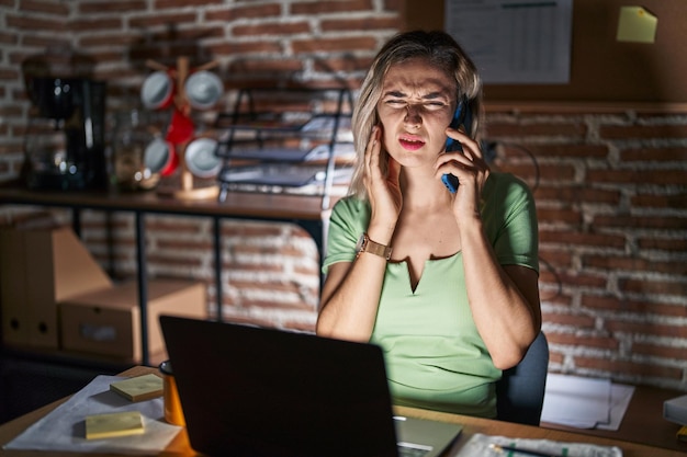 Kostenloses Foto junge schöne frau, die nachts im büro arbeitet und am telefon spricht und den mund mit der hand berührt, mit schmerzhaftem gesichtsausdruck wegen zahnschmerzen oder zahnerkrankungen an den zähnen. zahnarztkonzept