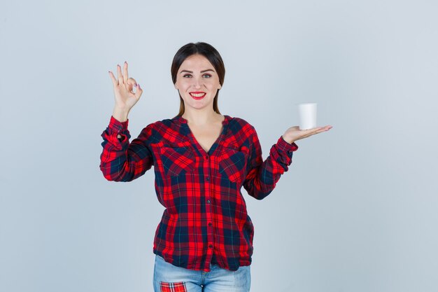 Junge schöne Frau, die Plastikglas hält, okayzeichen im lässigen Hemd, in den Jeans zeigt und glücklich schaut. Vorderansicht.