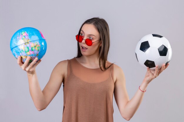 Junge schöne Frau, die rote Sonnenbrille hält, die Fußball und Globus betrachtet es überrascht, über weißem Hintergrund stehend