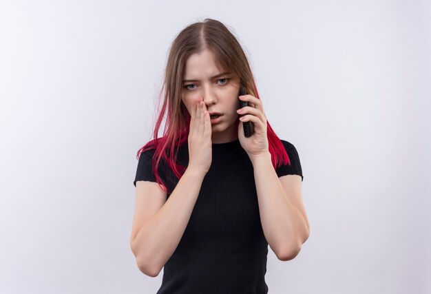 junge schöne Frau, die schwarzes T-Shirt trägt, flüstert am Telefon auf isolierter weißer Wand