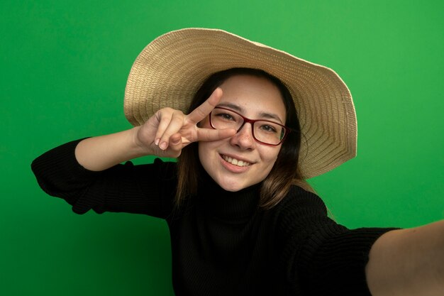 Junge schöne Frau, die Sommerhut in einem schwarzen Rollkragenpullover und in den Gläsern trägt, die Front glücklich und positiv lächelnd fröhlich machen V-Zeichen stehen über grüner Wand