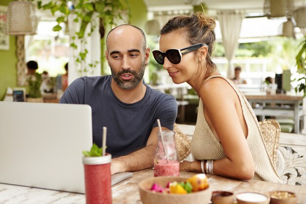 Junge schöne Frau, die Sonnenbrille und bärtigen Mann trägt, der an offener Terrasse sitzt und etwas auf ihrem allgemeinen Laptop-Computer beim Surfen im Internet unter Verwendung der drahtlosen Verbindung während des Mittagessens beobachtet