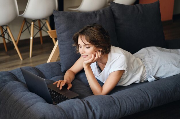 Junge schöne Frau, die verträumt auf dem Sofa liegt und im modernen Coworking Space am Laptop arbeitet