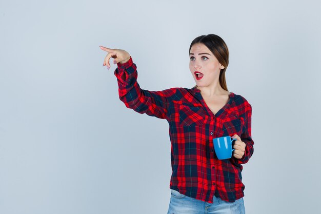 Junge schöne Frau, die weg zeigt, während sie eine Tasse in lässigem Hemd, Jeans hält und verwirrt aussieht, Vorderansicht.