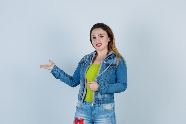 Junge schöne frau, die willkommensgeste in denim-outfit zeigt und fröhlich aussieht, vorderansicht.