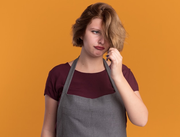 Kostenloses Foto junge schöne frau friseur in der schürze, die ihr haar unzufrieden steht, das über orange wand steht