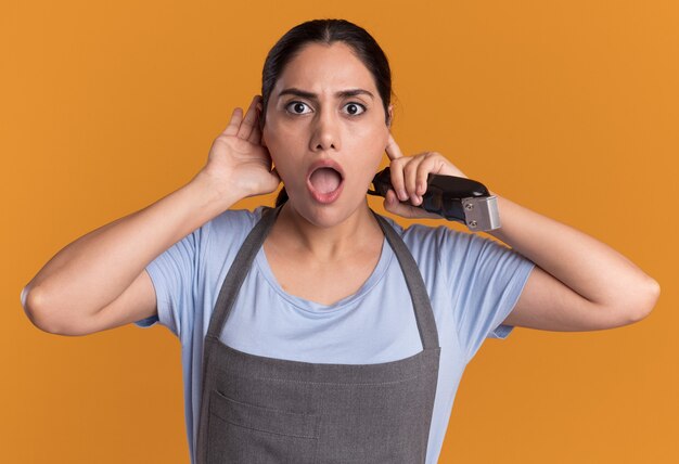 Junge schöne Frau Friseur in der Schürze hält Trimmer Hand über ihr Ohr schauend überrascht über orange Wand stehend