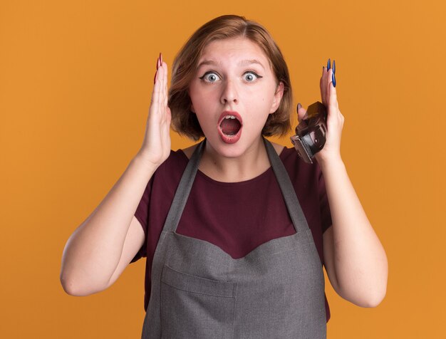 Junge schöne Frau Friseur in der Schürze hält Trimmer mit Blick auf die Front erstaunt und überrascht über orange Wand