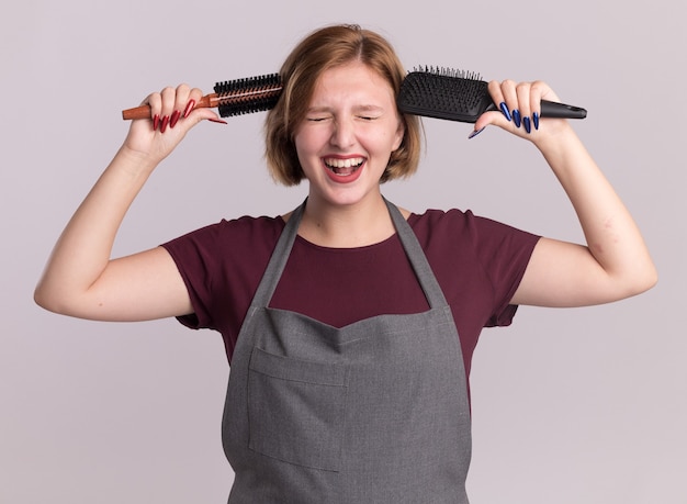 Junge schöne Frau Friseur in Schürze hält Haarbürsten in der Nähe ihres Kopfes lächelnd mit geschlossenen Augen über weiße Wand stehen