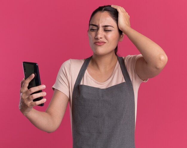 Junge schöne Frau Friseur in Schürze hält Smartphone betrachten es verwirrt und überrascht über rosa Wand stehen