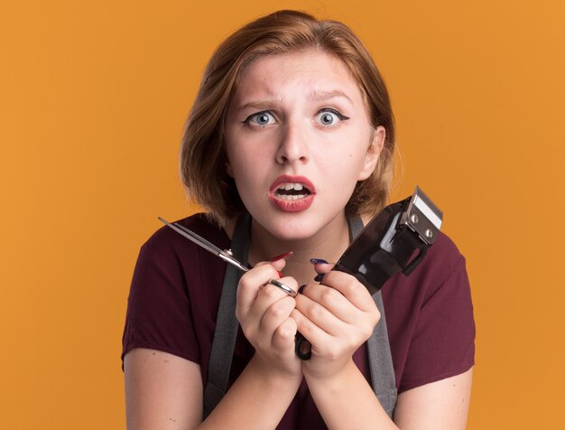 Kostenloses Foto junge schöne frau friseur in schürze hält trimmer und schere, die vorne besorgt über orange wand stehen