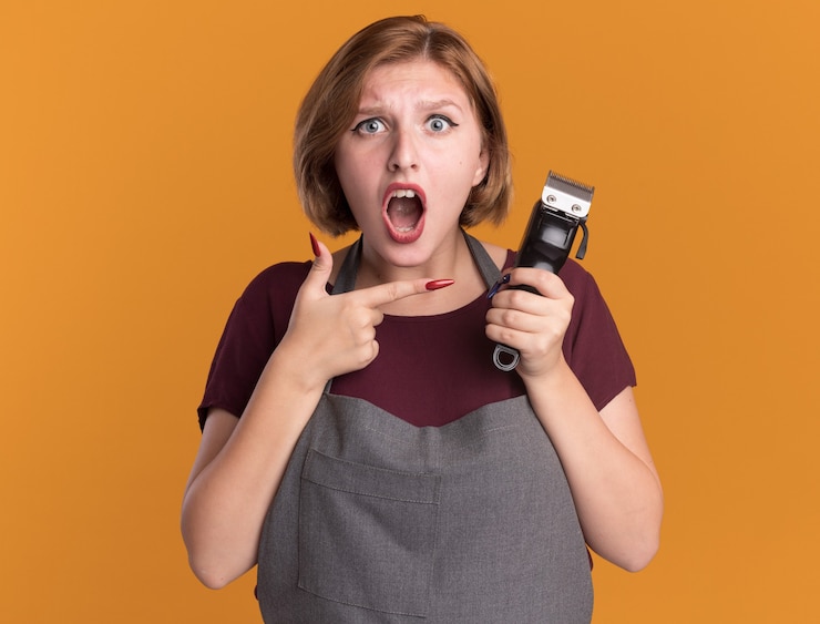 Kostenlose Foto junge schöne frau friseur in schürze hält trimmer zeigt mit zeigefinger darauf überrascht und verwirrt über orange wand stehen
