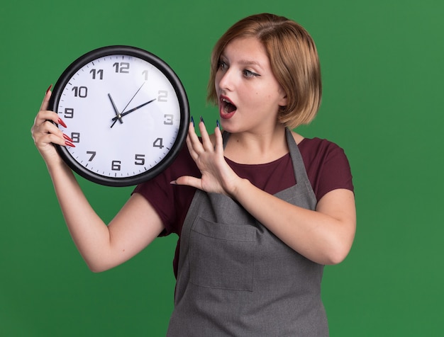 Junge schöne Frau Friseur in Schürze hält Wanduhr betrachten es erstaunt und überrascht über grüne Wand stehen